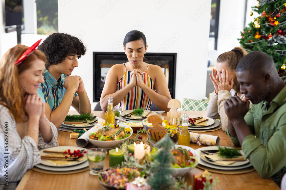 Happy diverse friends sitting at table and praying before dinner at christmas
