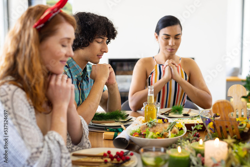 Happy diverse friends sitting at table and praying before dinner at christmas