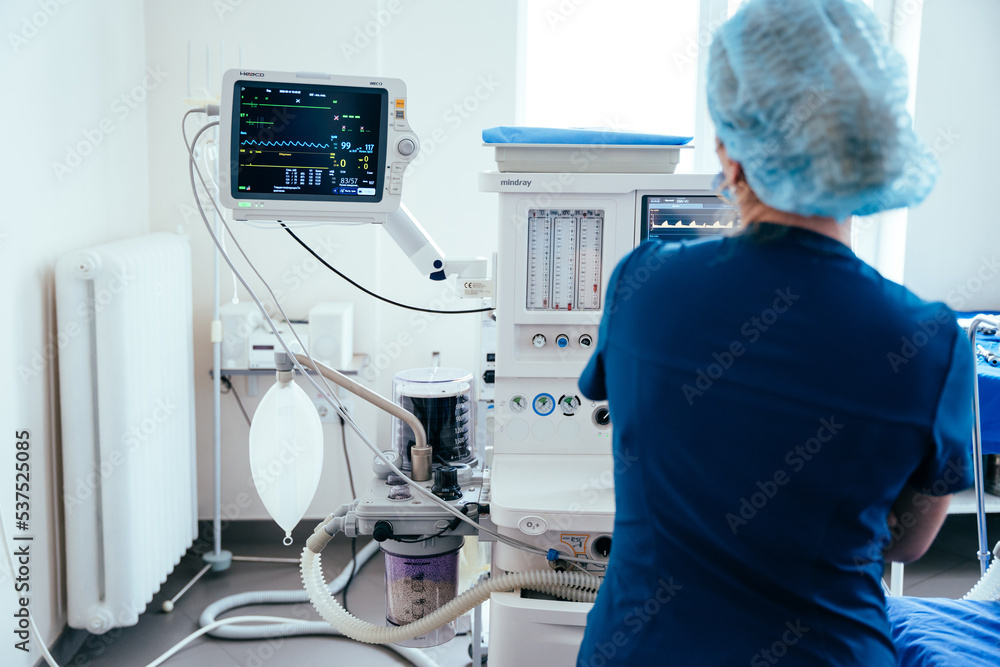 Fototapeta premium Rear view of female anaesthesiologist with monitor during operating in operation room. Modern equipment in clinic. Emergency room.