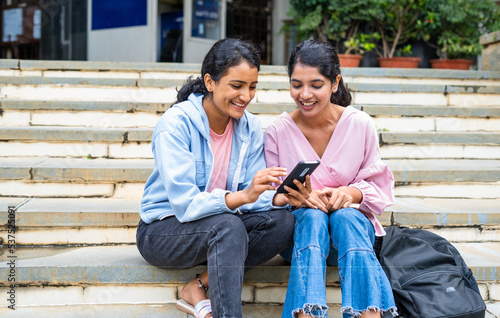 Happy smiling teenager girls checking result on mobile phone while sitting on campus at college - concept of technology, education and friendship.