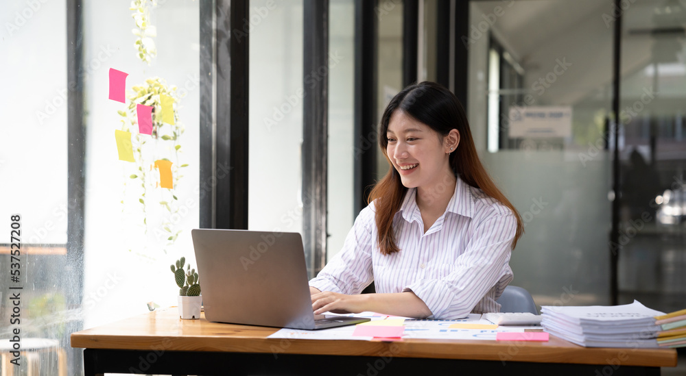 Happy young asian businesswoman sitting on her workplace in the office. Young woman working at laptop in the office. Happy young asian businesswoman sitting on her workplace in the office. Young woman working at laptop in the office.