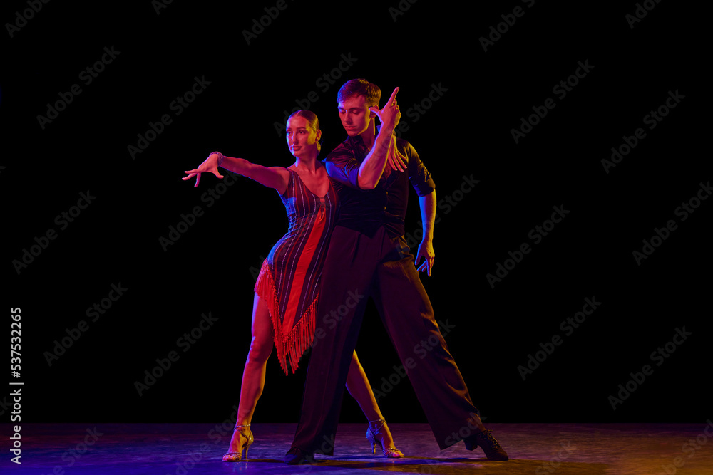 Fototapeta premium Stylish ballroom dancers couple in gorgeous outfits dancing in sensual pose on dark background in neon light. Concept of art, music, dance, emotions.