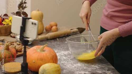 female blogger shoot video cooking pumpkin pie for thanksgiving family meal phone camera. unrecognizable hands whip eggs beat in bowl using metal whisk. record video for food vlog social media content photo