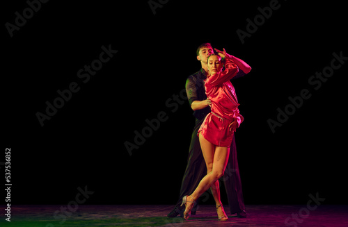 Full-length portrait of young beautiful man and woman dancing ballroom dance isolated over dark background in neon light. Concept of art, beauty, grace, action, emotions.
