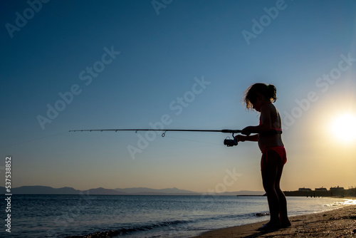 little girl is fishing with a fishing rod in the sea, under the sun. Silhouette.