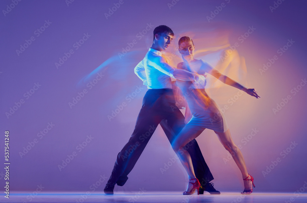 Young graceful dancers, flexible man and woman dancing ballroom dance isolated on gradient blue purple background in neon mixed light.