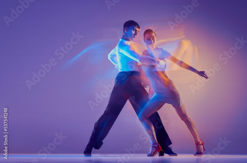Young graceful dancers, flexible man and woman dancing ballroom dance isolated on gradient blue purple background in neon mixed light. © master1305