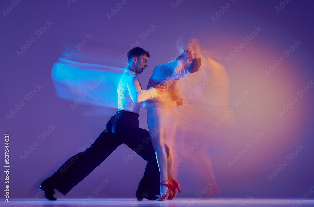 Fototapeta premium Art in motion. Flexible adorable ballroom dancers in stage attires dancing isolated on purple background in neon mixed light. Music, dance, emotions