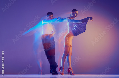 Young graceful dancers, flexible man and woman dancing ballroom dance isolated on gradient blue purple background in neon mixed light.