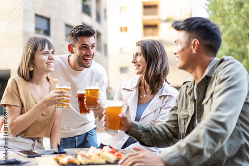 Multiracial friends drinking beer at brewery pub garden - Genuine friendship life style concept with guys and girls enjoying happy hour food together at open air bar dehor - Warm vivid filter