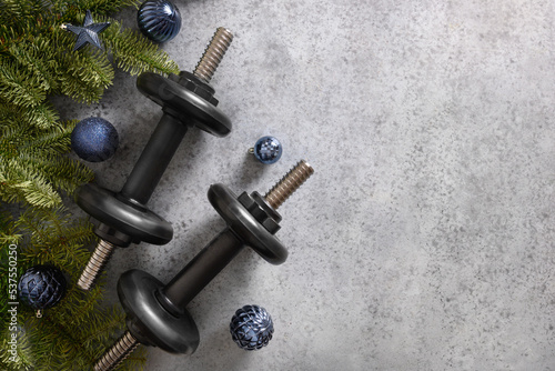 Christmas greeting card with two blue sports dumbbells, fir tree branches and gift on gray background. Top view with copy space. Fitness, sport and healthy concept. Xmas sport composition.