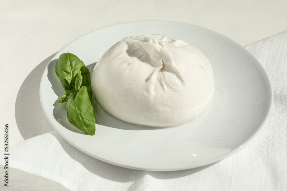 Traditional Italian, fresh, soft burrato ball of young mozzarella cheese with cream on a round white plate with basil greens on a light background. close-up