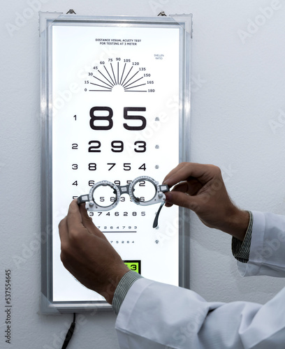 Eye doctor holding adjustable eye test trail frame with snellen number chart background in optical clinic, Ophthalmologist Glasses Concept photo