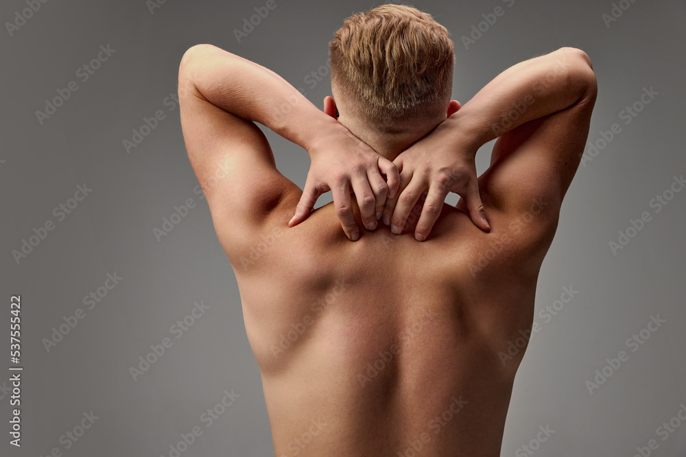 Obraz premium Portrait of young muscular man posing shirtless isolated over grey studio background. Pains in back, spine. Medical massage treatment