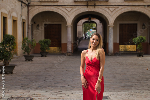 Young, attractive, blonde woman in an elegant red party dress smiling happily while strolling through the city. Concept beauty, fashion, elegance, luxury.