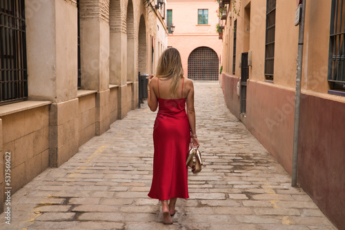 Young blonde woman, with her back turned, wearing an elegant red party dress and holding golden high heels in her hand, walking barefoot down a city alley. Concept beauty, fashion, elegance, luxury.