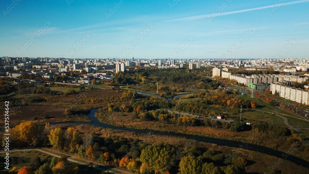 Autumn city park. Trees with colorful leaves. A winding bike path is visible between the trees. A river is flowing, city blocks are on the horizon. Autumn landscape. Aerial photography.