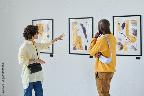 Young woman discussing paintings on wall together with African man while they visiting art gallery