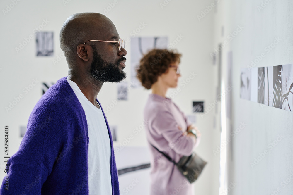 African man in eyeglasses looking at modern photos on the wall with woman standing in background African man in eyeglasses looking at modern photos on the wall with woman standing in background
