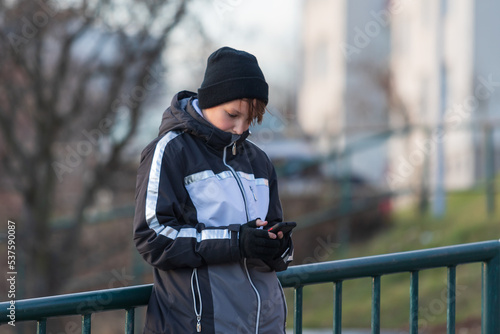 A young man about 12 years old in a winter jacket and cap is outside and holding a mobile phone.