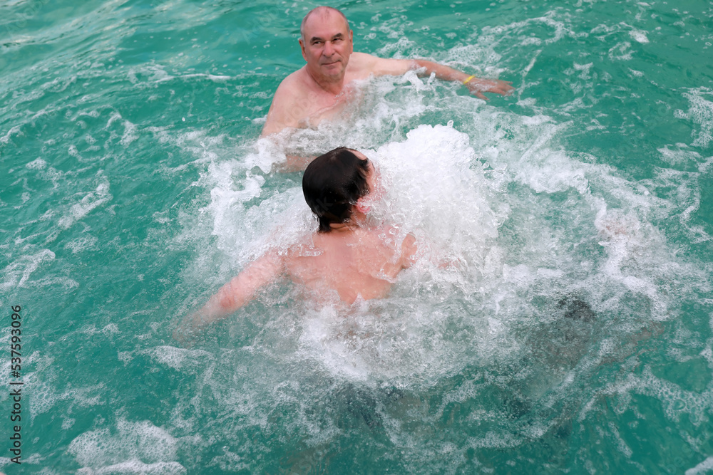 Grandfather with grandson get hydromassage in warm thermal pool