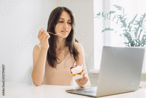 The concept of working at home. Young woman with a laptop in the kitchen  working with pleasure and eating delicious ice cream.