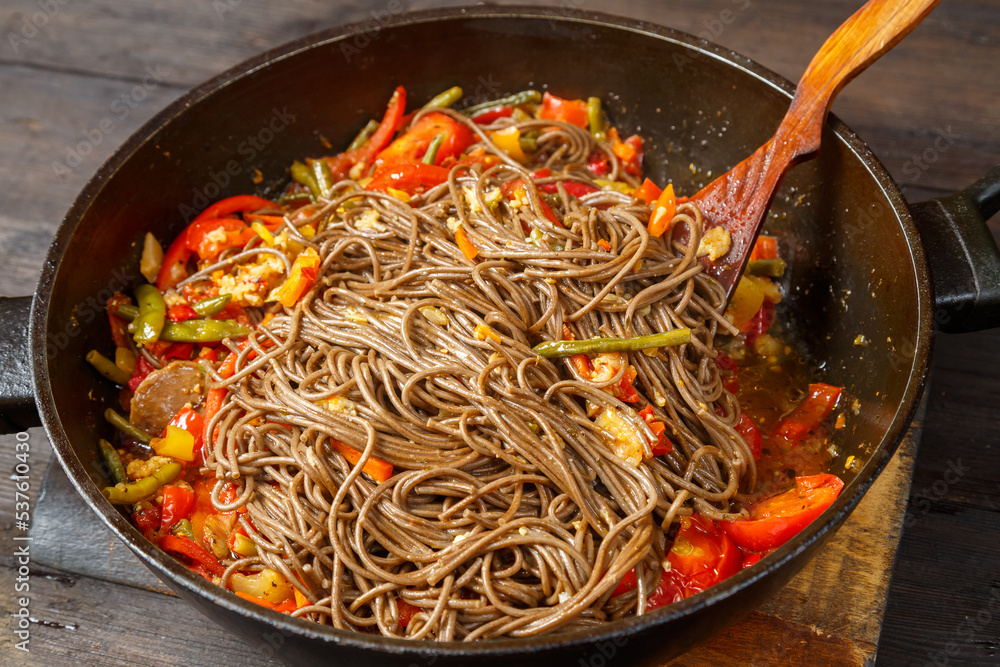 Soba with vegetables in soy sauce with spices in a pan with a spatula on a wooden stand on a wooden table. Soba with vegetables in soy sauce with spices in a pan with a spatula on a wooden stand on a wooden table.