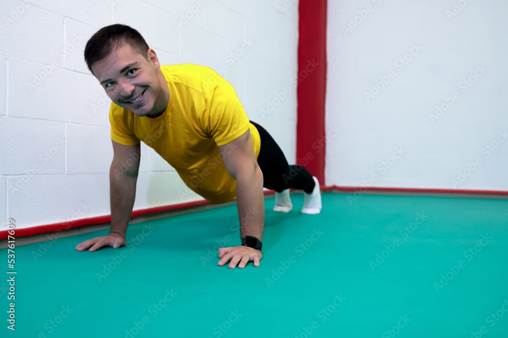 Portrait of a strong determined young man doing push-ups and looking at camera smiling. He is dressed in an yellow shirt and a black suit.