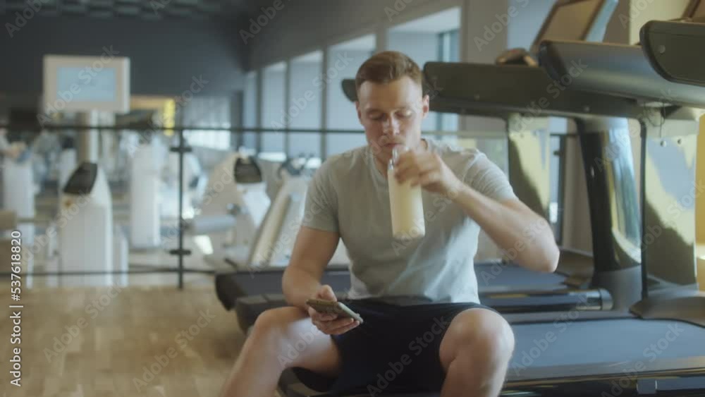 Muscular man in sportswear sitting on treadmill in gym, drinking water from bottle and browsing the web on smartphone while resting after workout Muscular man in sportswear sitting on treadmill in gym, drinking water from bottle and browsing the web on smartphone while resting after workout