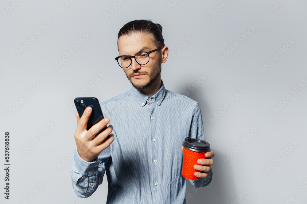 Studio portrait of young man looking in smartphone, holding a red paper cup for coffee takeaway in another hand, on white background. Wearing blue shirt and eyeglasses. Studio portrait of young man looking in smartphone, holding a red paper cup for coffee takeaway in another hand, on white background. Wearing blue shirt and eyeglasses.