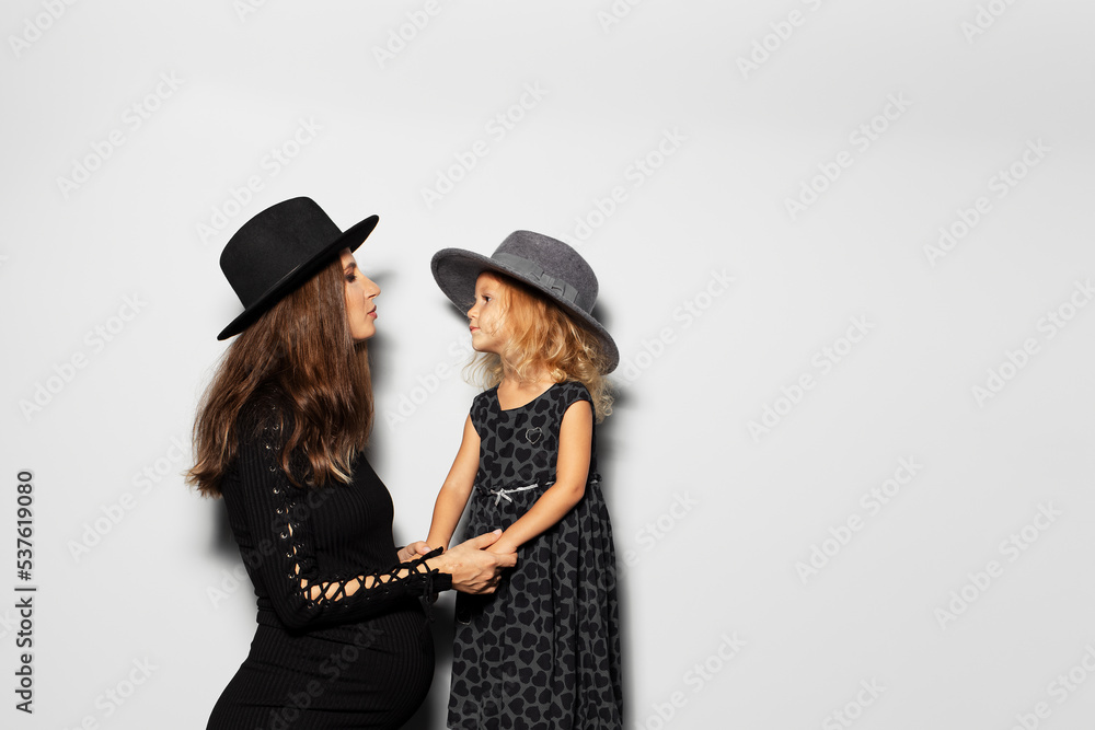 Studio portrait of pregnant smiling woman and child girl looking each ever, wearing hats on white background. Mother and daughter in black. Studio portrait of pregnant smiling woman and child girl looking each ever, wearing hats on white background. Mother and daughter in black.