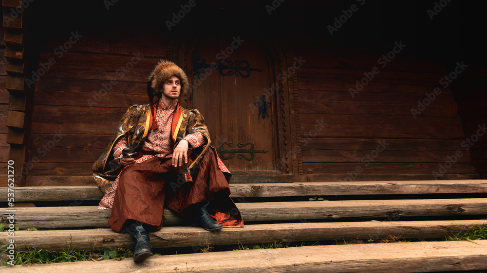 Pensive young ukrainian cossack dressed traditional clothing holding saber in his hand sitting on the steps and looking into the distance on background of old wooden door.