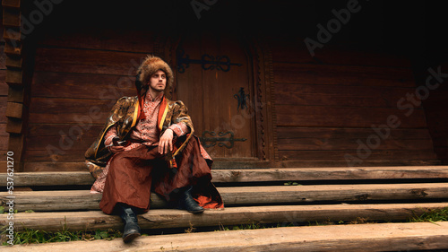 Obraz na płótnie Pensive young ukrainian cossack dressed traditional clothing holding saber in his hand sitting on the steps and looking into the distance on background of old wooden door. w salonie