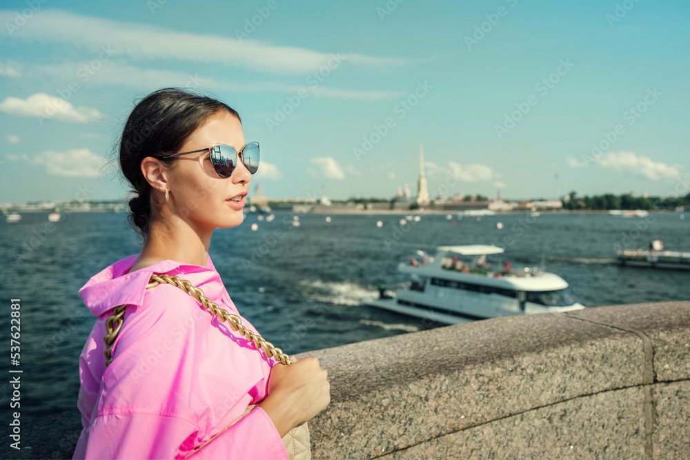 Young lady wearing sunglasses and pink clothing spending time outdoor walking at urban street against river with boats Young lady wearing sunglasses and pink clothing spending time outdoor walking at urban street against river with boats