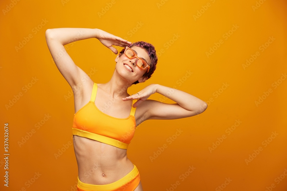 Fototapeta premium Young woman in a bright yellow fitness tracksuit with an open belly and sunglasses on an orange background smiling