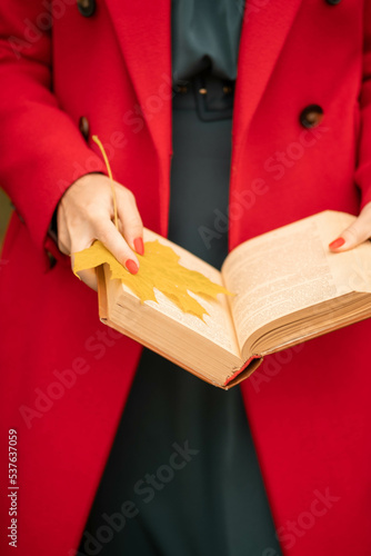 A European girl in a red coat holds an open book with a yellow maple leaf in her hands