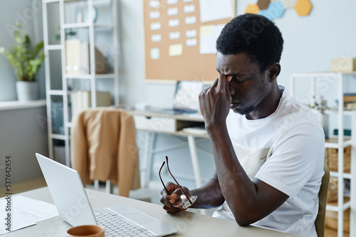 Portrait of frustrated black man taking off glasses at workplace and suffering from headache