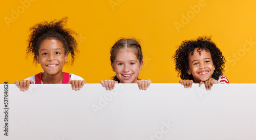 Group of cheerful happy multinational children with blank white poster on  yellow background