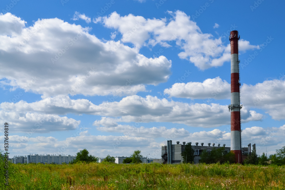 Tall striped red and white chimney of a boiler house against the background of white clouds in the blue sky