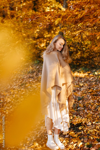 A woman in a brown poncho enjoys autumn. The girl is standing in the middle of the yellow leaves of the trees.