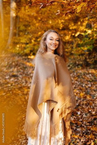 A woman in a brown poncho enjoys autumn. The girl is standing in the middle of the yellow leaves of the trees.