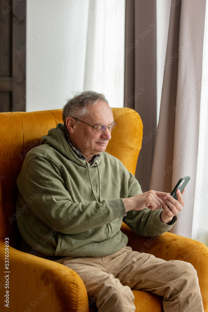 Senior male sits in armchair at home and using mobile phone