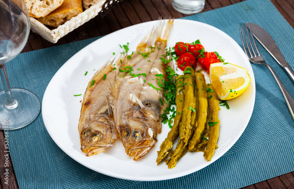 Whole oven baked rooster fish with marinated asparagus and tomatoes on white plate