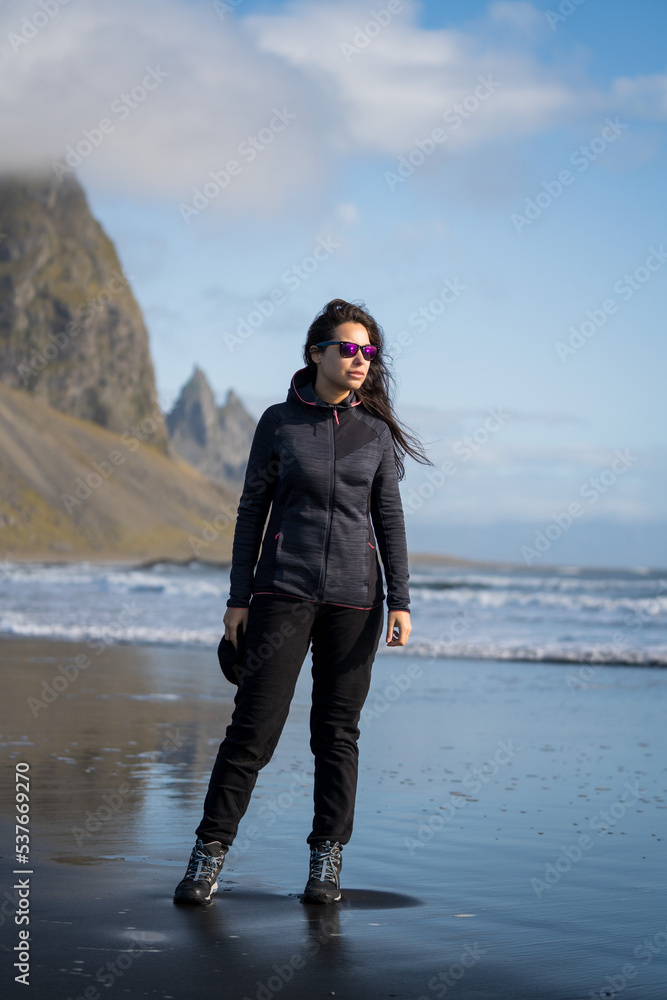 adventurous woman with cap and sunglasses walking on the black volcanic sand beach at the seashore with mountains in autumn winter adventurous woman with cap and sunglasses walking on the black volcanic sand beach at the seashore with mountains in autumn winter