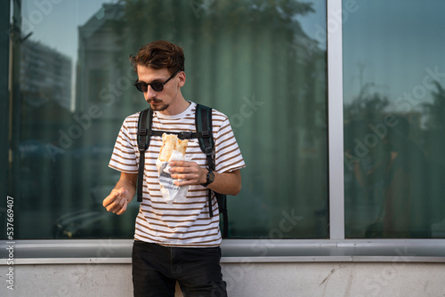 One man young adult modern caucasian male in the city in sunny day stand and eat sandwich fast food concept urban life copy space tourist eating in front of building wall real person portrait