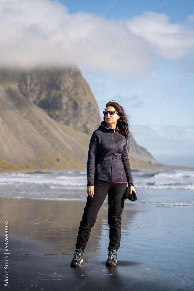 adventurous woman with cap and sunglasses walking on the black volcanic sand beach at the seashore with mountains in autumn winter adventurous woman with cap and sunglasses walking on the black volcanic sand beach at the seashore with mountains in autumn winter