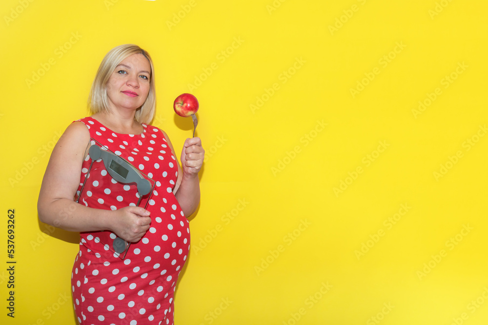 pregnant woman in a red polka dot dress with scales and an apple on a fork on a yellow background with copy space pregnant woman in a red polka dot dress with scales and an apple on a fork on a yellow background with copy space