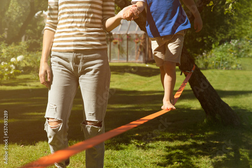 Mother holding kids hand walking on slackline training balance with bear foot on a warm sunny summer day in a green garden. Healthy living concept. Balance and help concept.