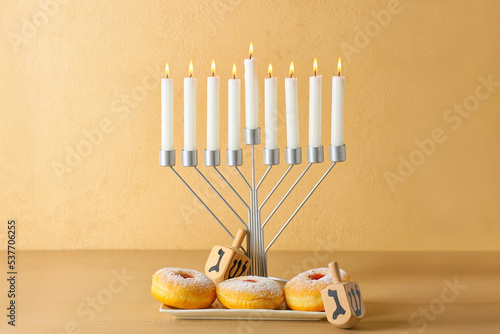 Menorah with candles, dreidels and delicious donuts for Hanukkah celebration on wooden table against color background