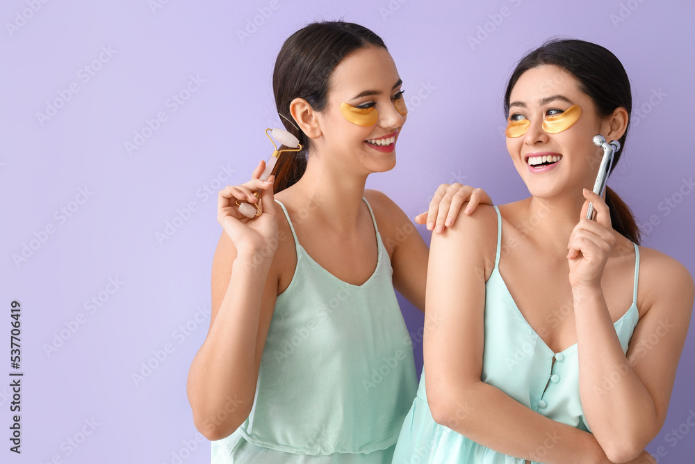 Beautiful young women with under-eye patches and facial massage tools on lilac background Beautiful young women with under-eye patches and facial massage tools on lilac background